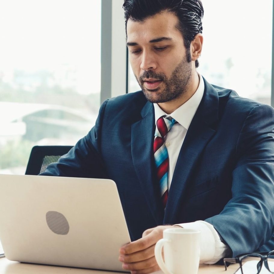 Skill Loop Collective Service Page Man in a suitWorking on a laptop at a desk with a coffee mug and city view behind him.